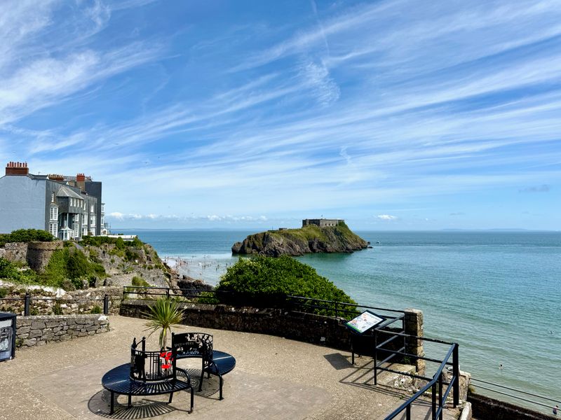 A small patio overlooks the water with some buildings over the left. Just off the coast is a rocky island with a building on it.