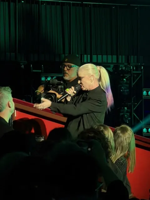 Shirley Manson leaning over the balcony railing into the crowd, singing with a handheld microphone, with a camera operator filming behind her.