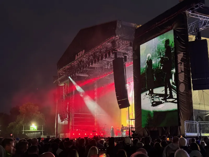 A lively outdoor concert scene at night, featuring a large stage with colorful lights and a captivated audience.