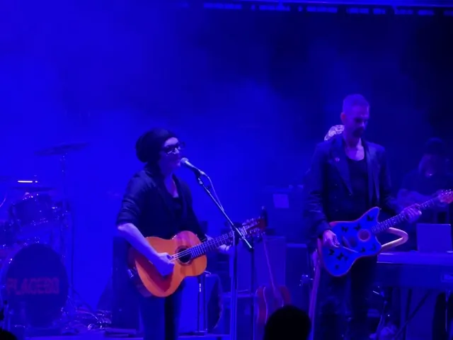 Brian Molko playing an acoustic guitar and singing into a microphone, wearing a beanie and glasses, with a guitarist playing a blue electric guitar to his right under deep blue stage lighting. The Placebo drum kit is visible in the background.