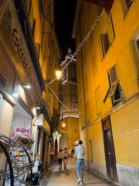 Looking down the narrow streets of Vieux Nice at night. The buildings are yellow and aged and a woman is sitting outside a shop. At the end of the street is a large cathedral belltower.