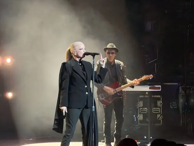 Shirley Manson singing into a microphone in a black blazer, with a guitarist in a hat and red semi-hollow guitar performing behind her in smoky stage lighting.