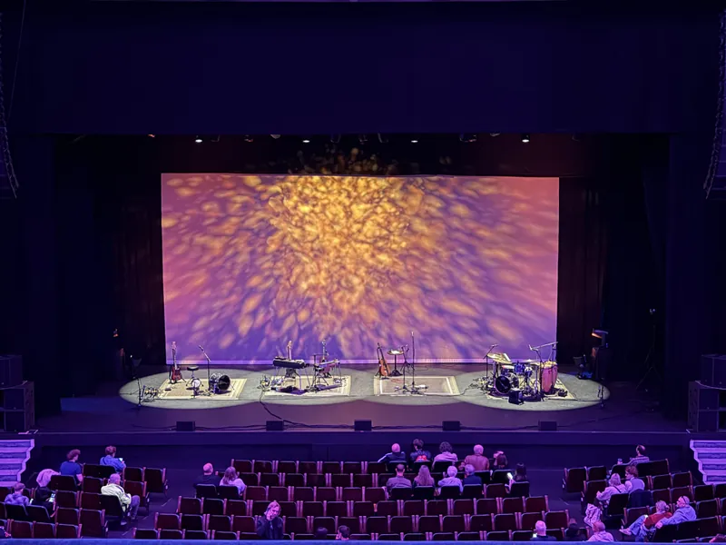 Another balcony view of the empty stage, instruments and amps waiting on four spotlit rugs beneath a glowing amber and purple light pattern, with the audience filling in below.