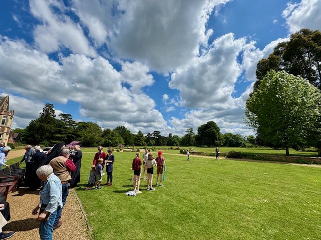 Outside on a green lawn people milling about and trees in the distance. Huge white fluffy clouds hang over head with patches of bright blue sky.