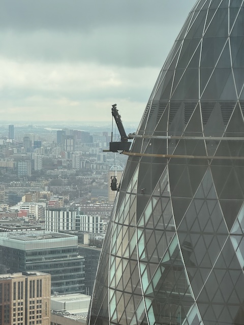 A crane attached to the side of the gherkin in London with someone hanging down washing windows