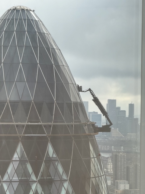 A crane attached to the side of the gherkin in London that is lifting someone up to the top of the building to wash windows