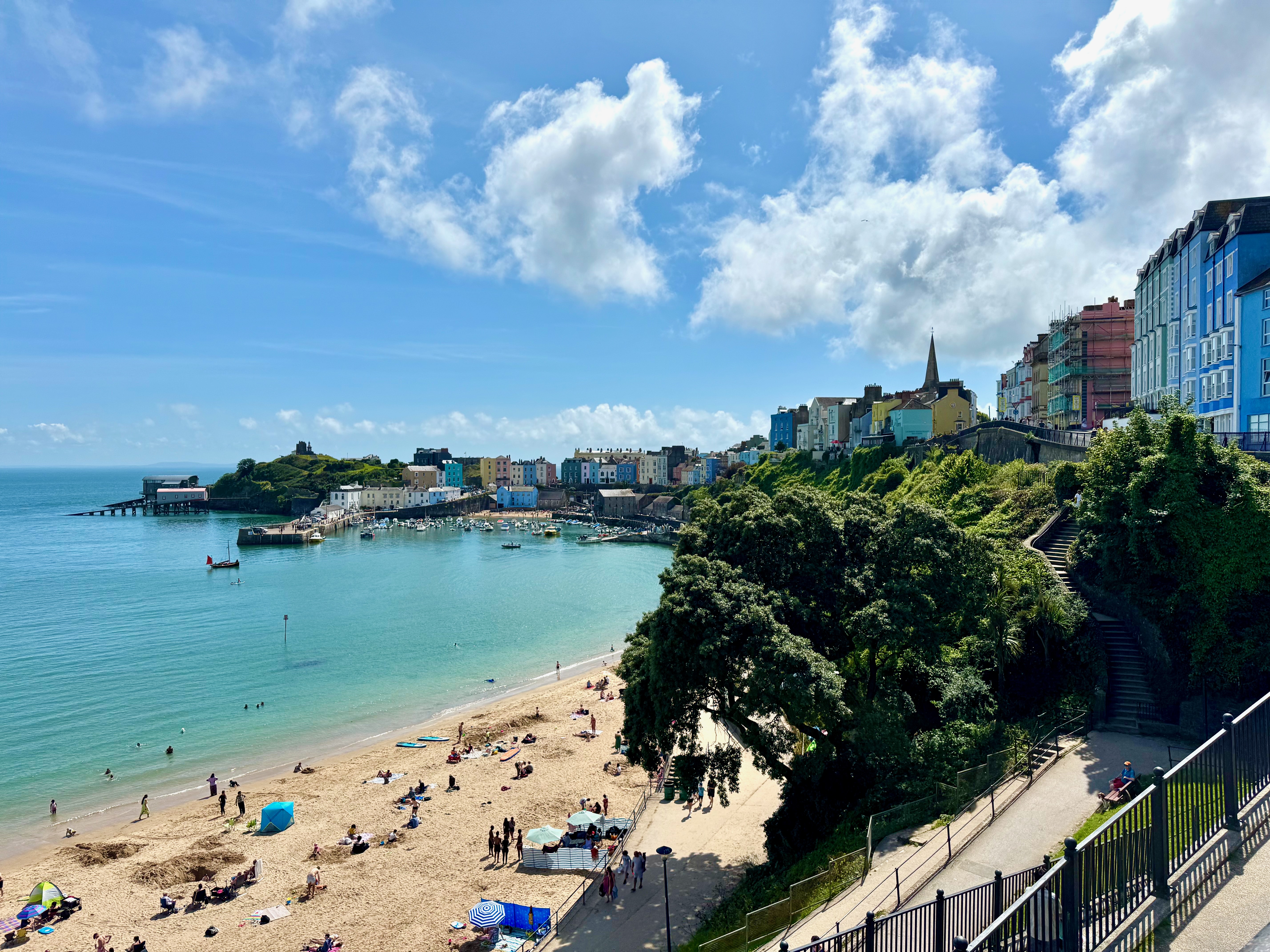 Looking down on the beach towards the town of Tenby. There is a harbour in the middle with smaller fishing and pleasure boats. Colourful buildings line the cliff that sits above the beach