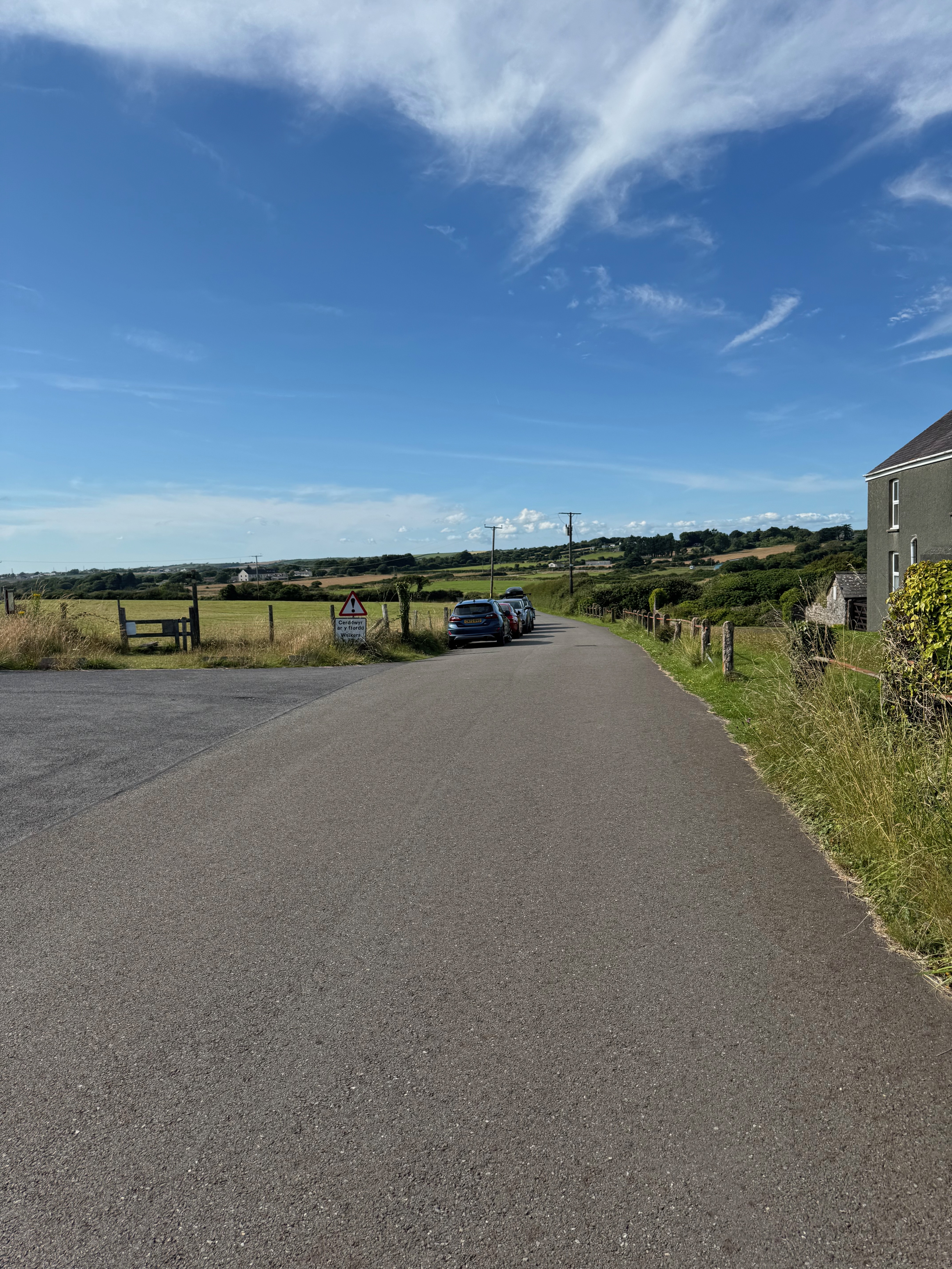 Looking down a paved country road. Cars parked on the side of the road to the left.