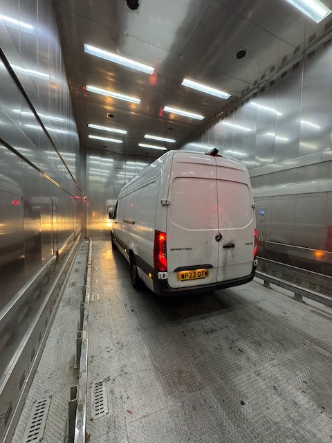 A white panel van inside a giant chrome box, which is actually a lift for large trucks.