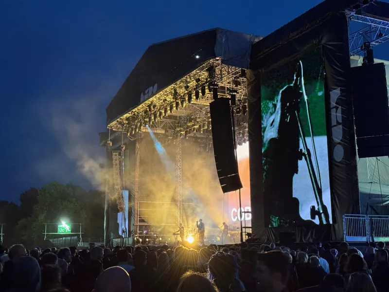 Large outdoor concert stage with a crowd of people, illuminated by spotlights and a large video screen displaying a performer.