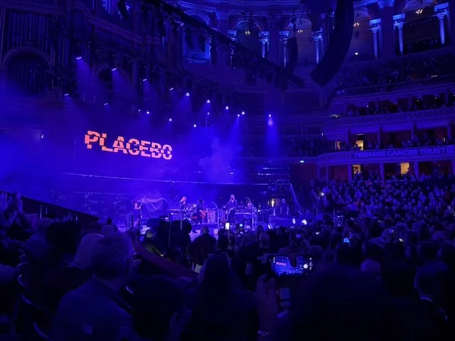 Wide shot of the Royal Albert Hall with "PLACEBO" in orange letters on the LED screen, the band performing on stage bathed in blue light, with a packed crowd and a "Teenage Cancer Trust" banner on the balcony.