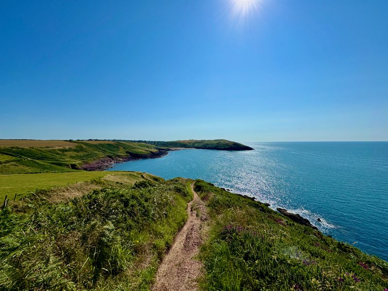 Manorbier Beach in the distance with the blue water of the bay in the foreground and along the right. The sun is shining bright in a cloudless sky