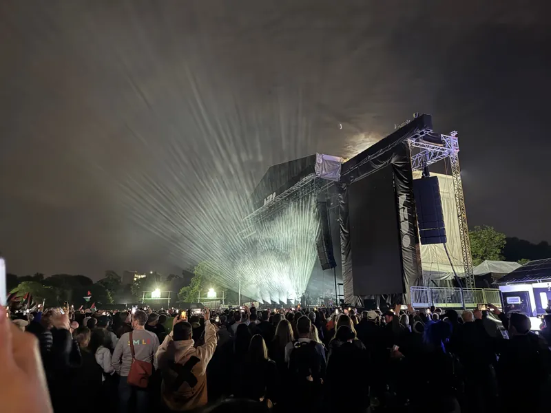 Outdoor concert at night with a large crowd facing an illuminated stage. Lights create beams in the cloudy sky, adding drama and excitement.