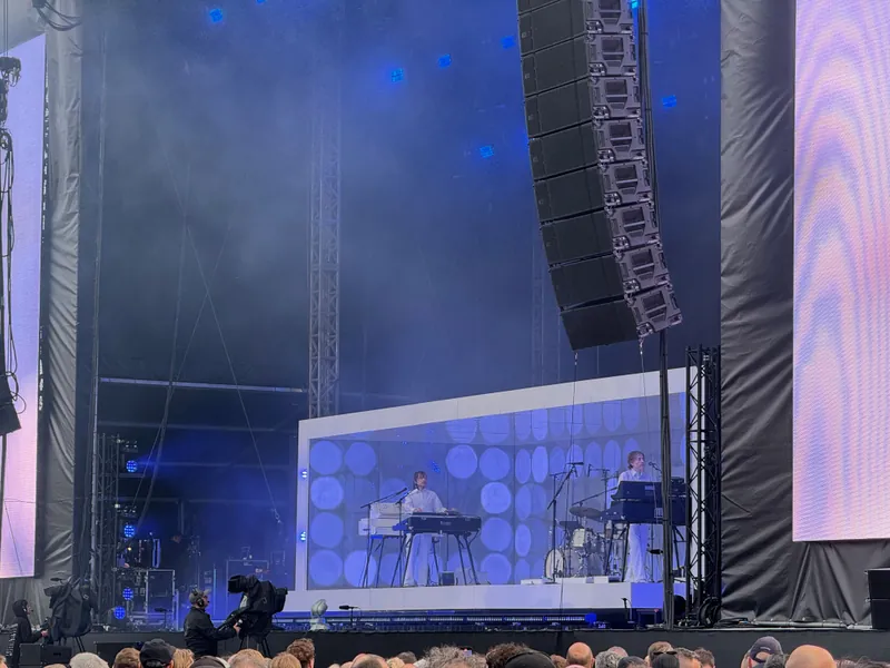 Two musicians playing keyboards on a stage, with large sound equipment and a large screen backdrop.