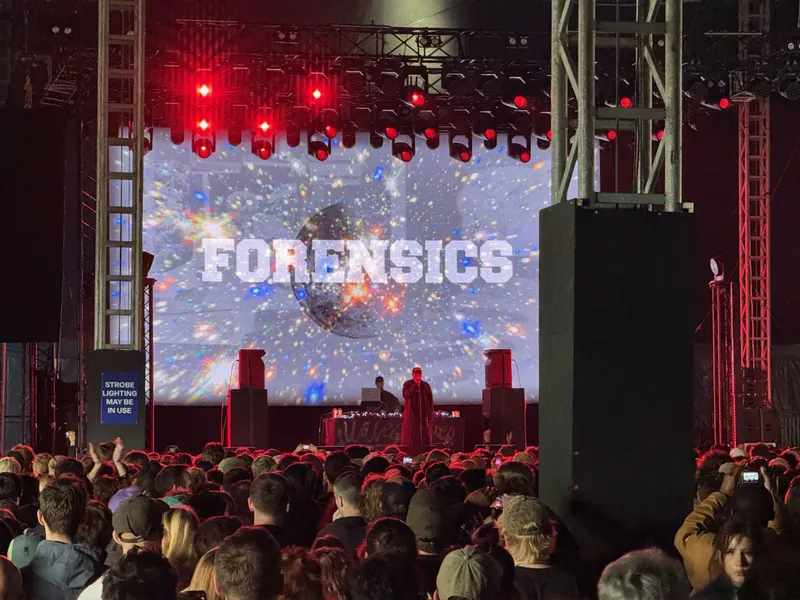 A crowd faces a stage with a DJ performing in front of a large screen displaying the word "FORENSICS" and a space-themed background, under red stage lights.