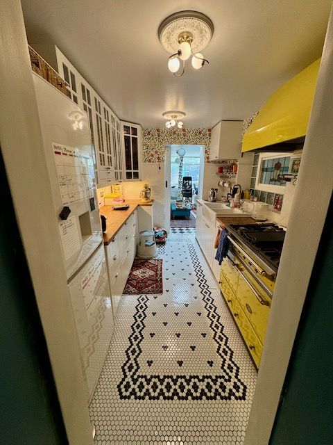 wide angle of a mostly white kitchen with wooden counter tops and a yellow stove. There is a black and white pattern in tile on the floor