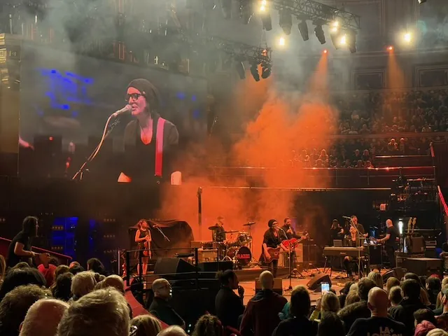 Wide shot of the Royal Albert Hall stage with orange smoke billowing across it, a large screen showing a close-up of the singer, and the full band performing to a packed crowd.