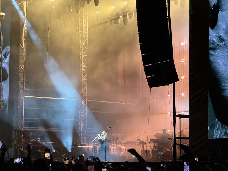 A performer sings passionately on stage, illuminated by dramatic lighting and surrounded by a captivated audience with smartphones raised.
