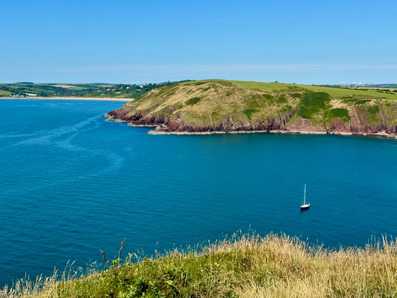 Looking down into a blue bay with a lone sail boat anchored in the bay. Off in the distance there is another long beach stretching along the coastline.