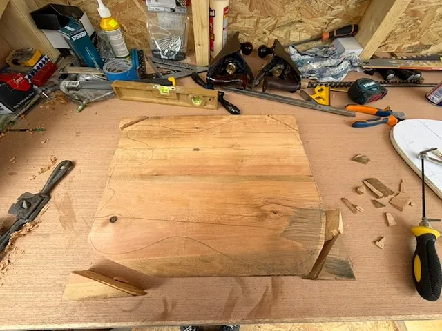 A wooden board on a workshop workbench, with pencil outlines sketched on its surface suggesting a shape being marked for cutting. The board shows natural wood grain and some knots. Surrounding it are various woodworking tools including a spirit level, screwdrivers, pliers, clamps, chisels, and wood shavings.
