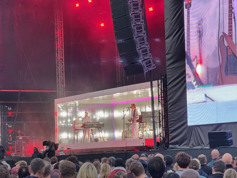 Band performing on a large stage with a glass-enclosed performance area, surrounded by a crowd of concertgoers.