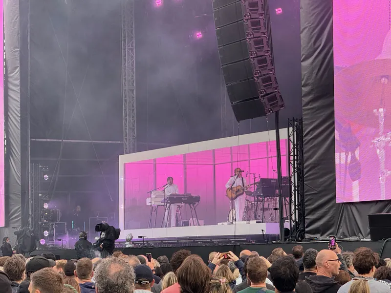 Concert stage with musicians playing keyboards and acoustic guitar, large crowd in the foreground.