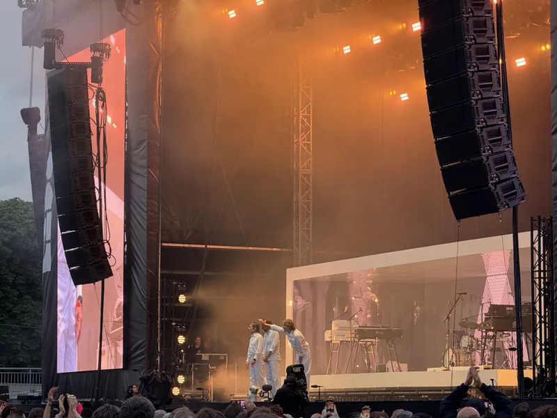 Three musicians in white outfits perform on a stage with large sound speakers and a large screen.