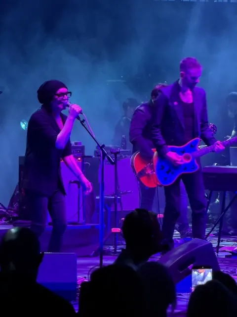 Close-up of Brian Molko singing into a microphone, wearing a beanie and glasses, with a guitarist playing a blue electric guitar beside him under purple stage lighting.