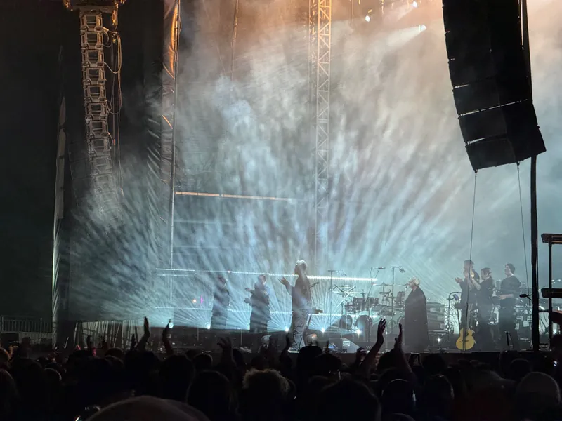 Concert scene with bright lights radiating behind a singer and band on stage. Crowd in foreground, cheering and clapping. Energetic atmosphere.