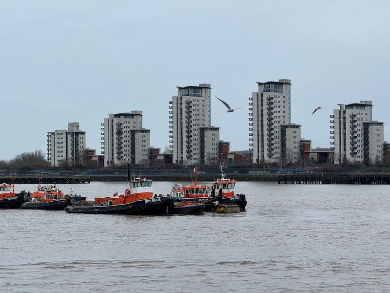 Some tug boats parked in  the middle of the Thames river with apartment buildings on the far shore